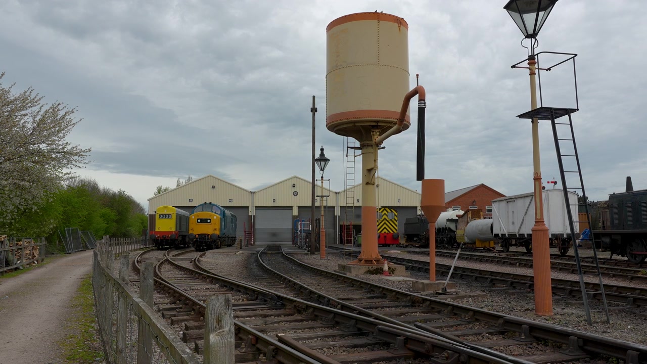 Un'inquadratura statica di locomotive diesel d'epoca parcheggiate vicino ai depositi locomotive e alla torre dell'acqua in un sito di conservazione ferroviaria sotto un cielo coperto.