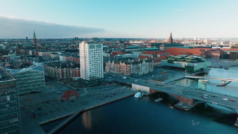 Bewegte-Drohnenaufnahme-Der-Skyline-Der-Innenstadt-Von-Kopenhagen-In-Dänemark-Am-Fluss-Tryggevælde-Mit-Einer-Brücke-In-Der-Rechten-Ecke