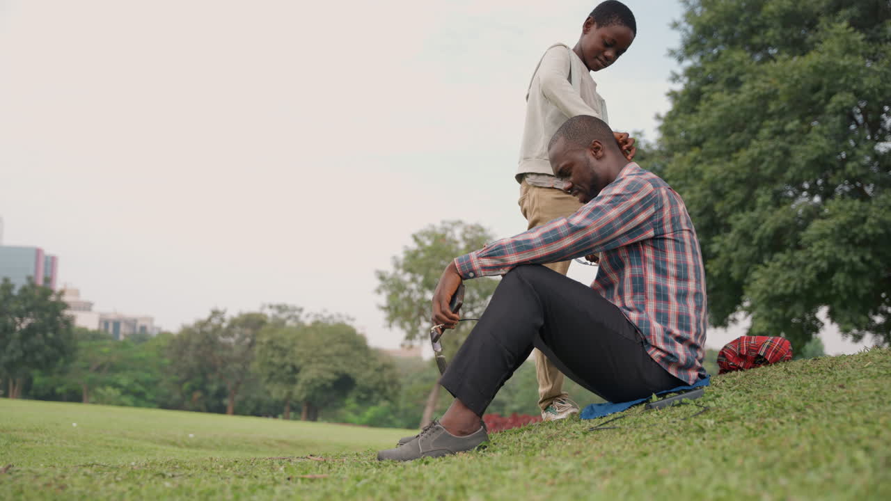 Schwarzer Vater und Sohn im Park teilen einen unbeschwerten Picknick-Moment. Der Sohn neckt den Vater, während beide lachen. Lässiges kariertes Hemd, roter Rucksack in der Nähe, warme Sonne. Eine spielerische Bindung und optimistische Verbindung.