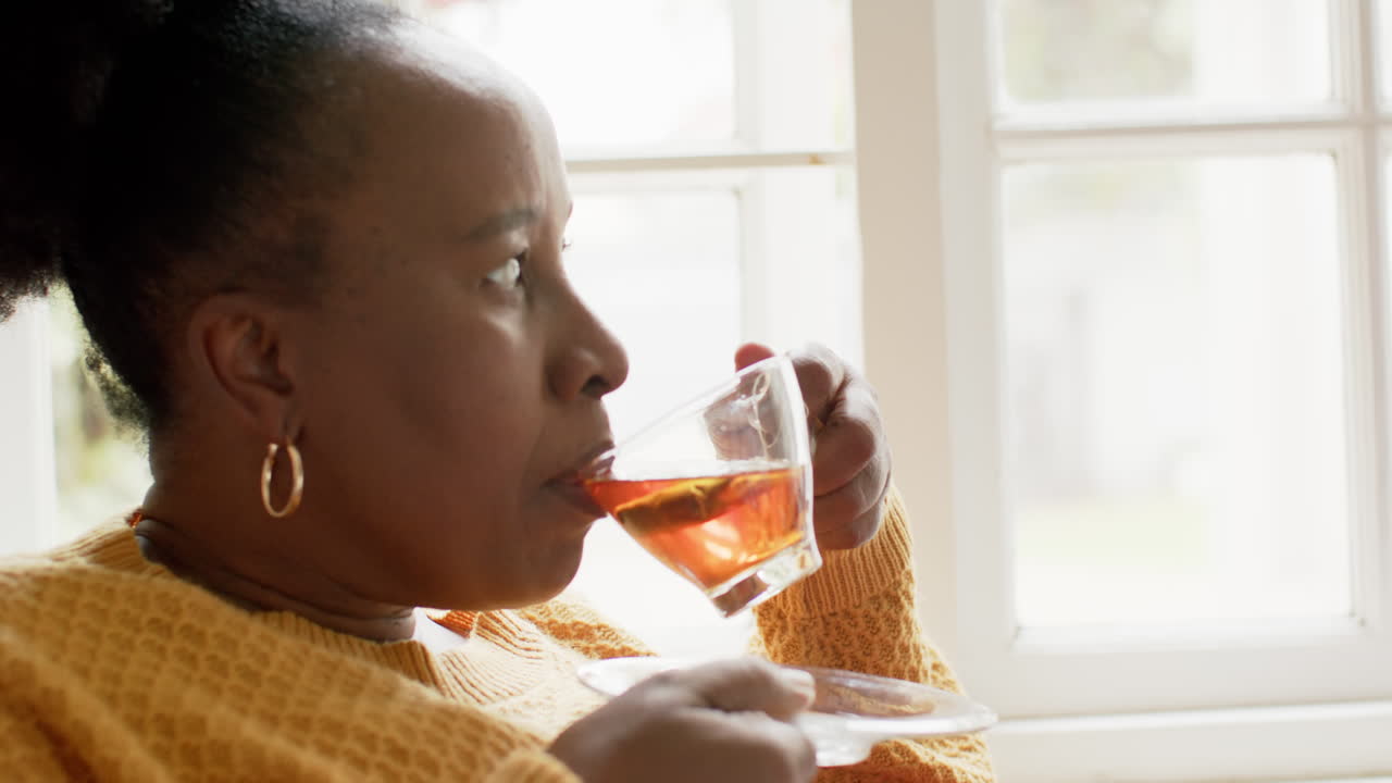 Happy african american senior woman drinking tea and looking ahead in sunny room, slow motion