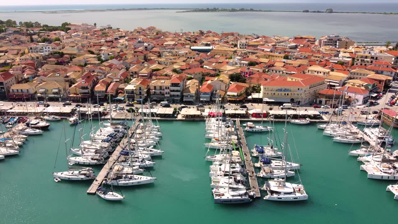 Vista aerea della Marina di Lefkas, Lefkada, isola ionica in Grecia. Barche a vela nel porto, pedoni e traffico che si muovono lungo il lungomare accanto al centro storico color terracotta, scena estiva mediterranea.
