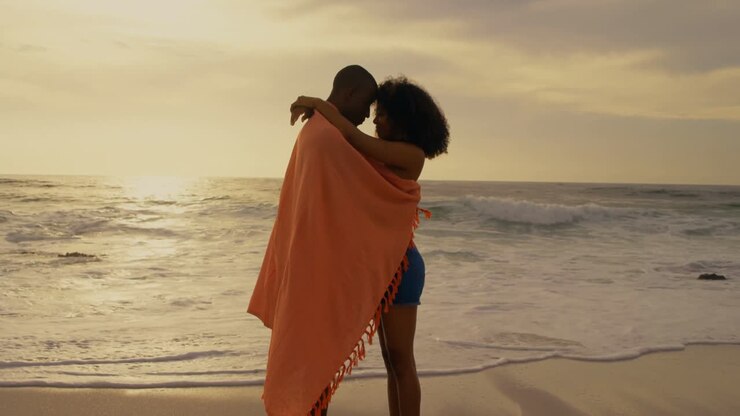 Vista laterale di coppia afroamericana avvolta in una coperta sulla spiaggia 4K