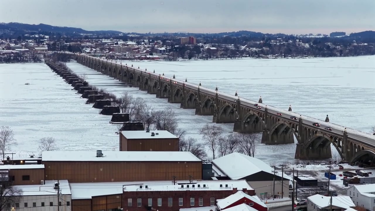 Vista aerea ascendente di Wrightsville, Pennsylvania, USA, con tetti coperti di neve, auto, un ponte storico e un fiume ghiacciato durante una leggera nevicata invernale. Atmosfera natalizia pittoresca di una piccola città in A.