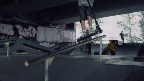 millennial man jumping on rail with scooter at urban city skate park.