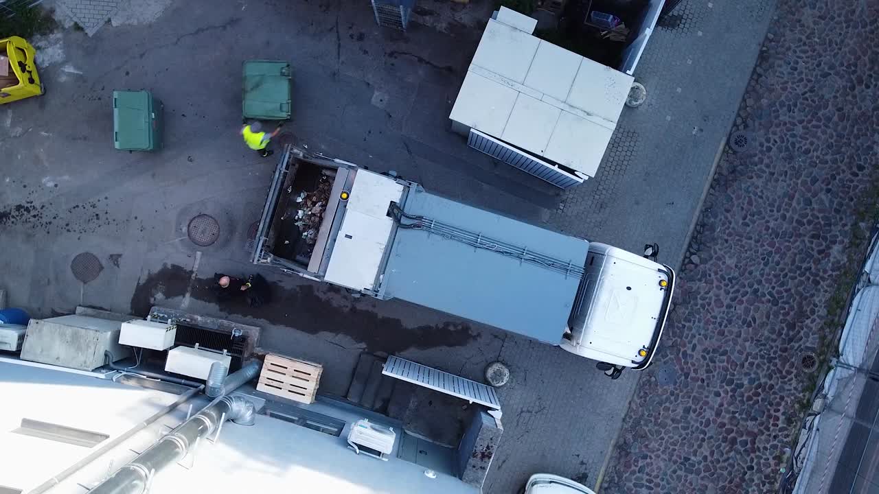 Vista aerea di un camion della spazzatura che carica i rifiuti automaticamente, persone che spostano i cassonetti, ripresa drone grandangolare a volo d'uccello che ruota lentamente a sinistra.