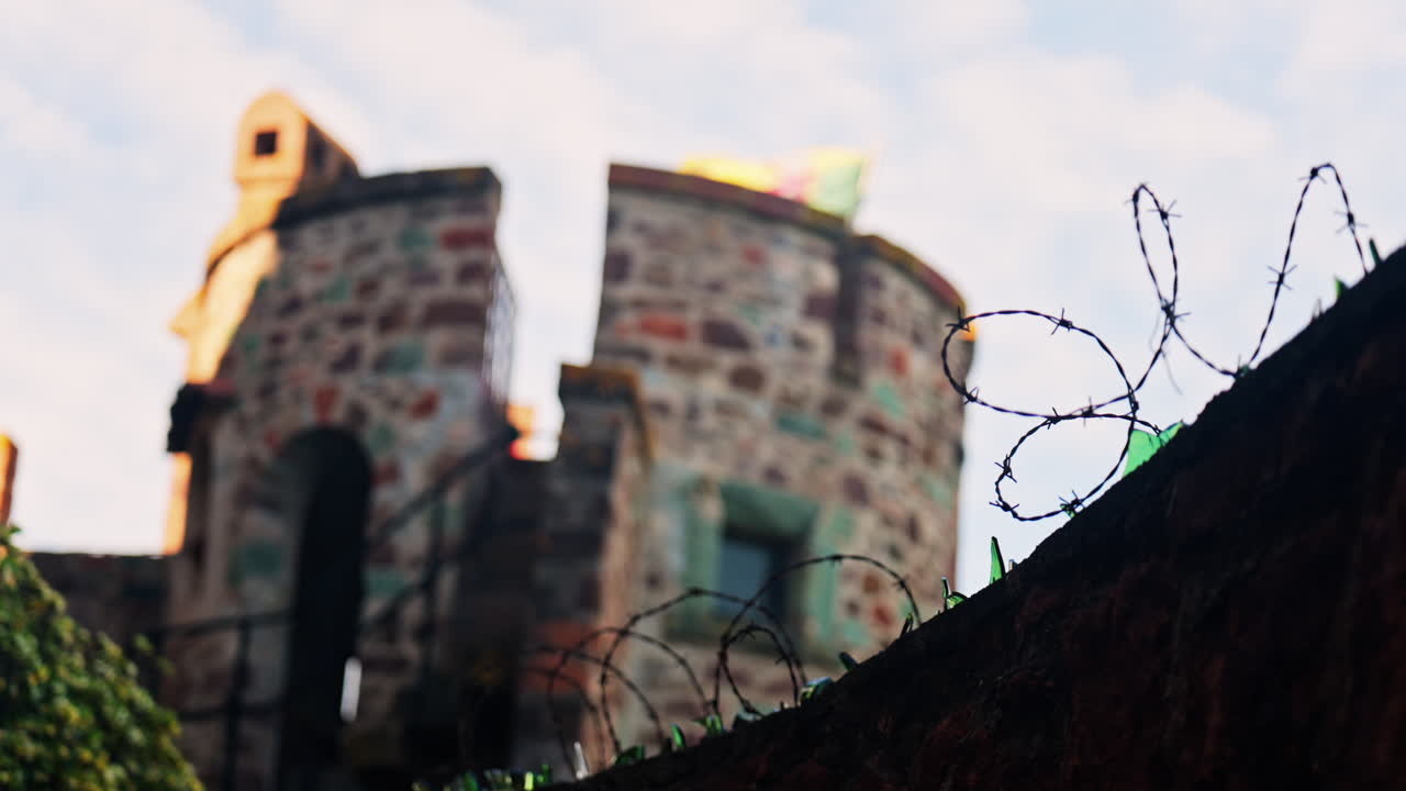 Close up of multiple broken glass shards and barbed wire loops along the edge of a brick wall with a blurred view of the Chateau de la Napoule Castle in Mandelieu-La Napoule, France