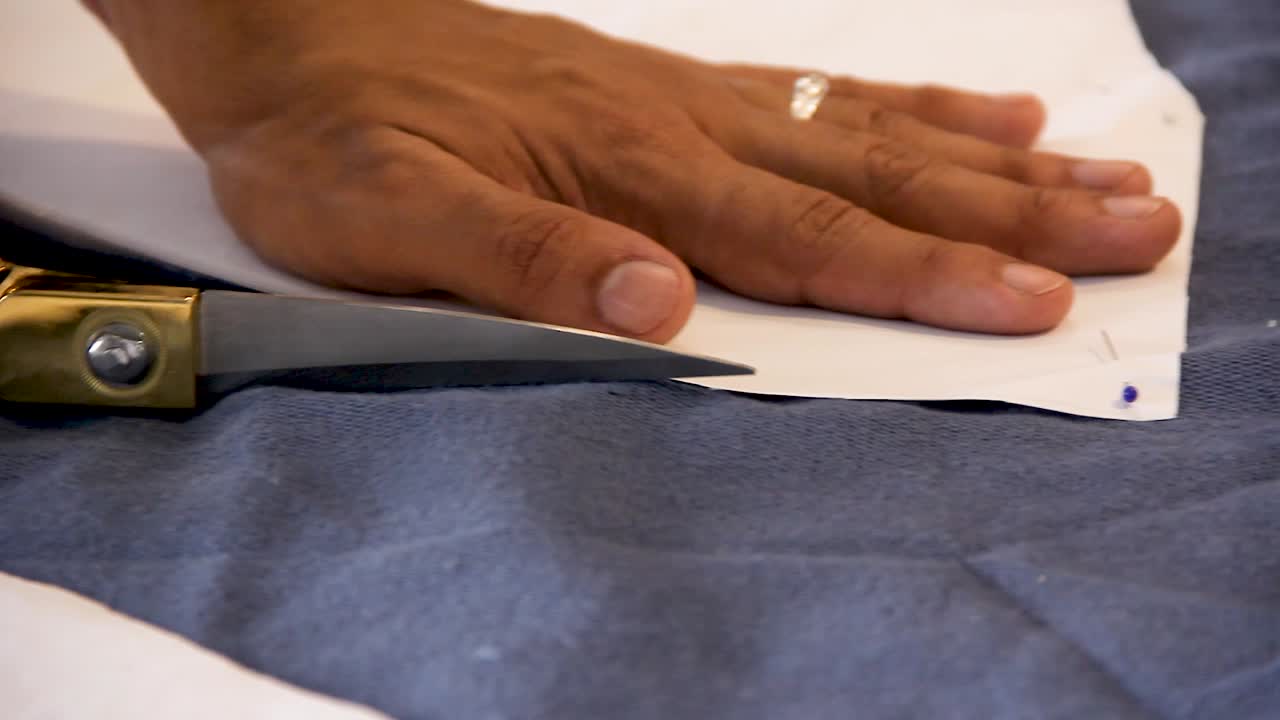 Macro close up of male hands cutting a blue fabric with golden scissors from a sewing pattern attached with colorful pins - shallow depth of field handheld interior shot natural light