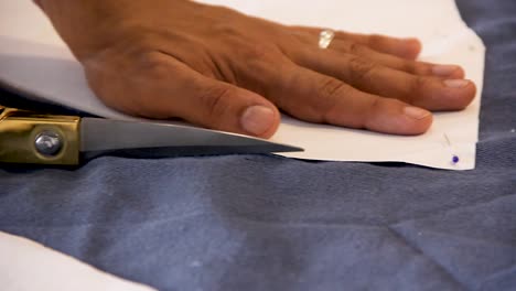 Macro-close-up-of-male-hands-cutting-a-blue-fabric-with-golden-scissors-from-a-sewing-pattern-attached-with-colorful-pins---shallow-depth-of-field-handheld-interior-shot-natural-light