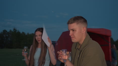 man sipping champagne from glass while woman waves certificate to swat buzzing insects around them on grassy field at dusk creating playful protective moment after scenic hot air balloon flight