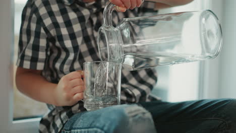 close up of kid thoughtfully seated indoors wearing plaid shirt carefully pouring clear water from large glass jar into transparent cup resting on denim jeans