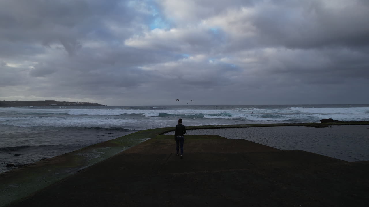 toma aérea de una mujer de mediana edad que camina hacia la costa donde rompen las olas y durante la puesta de sol, en la playa de boca barranco en la ciudad de gáldar, en la isla de gran canaria