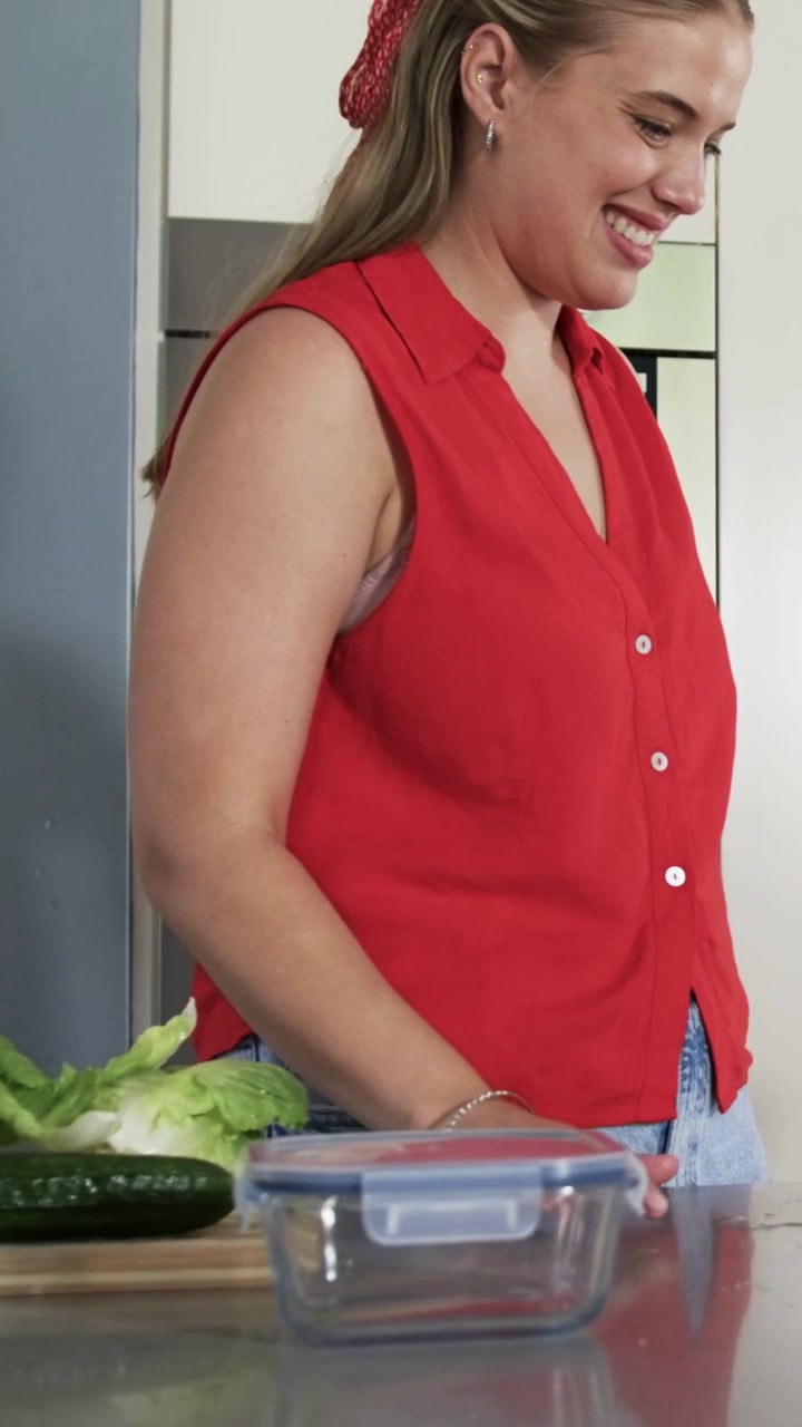 Video verticale: Donna con top rosso nota un cetriolo, afferra la lattuga e la mette in una scatola sul bancone della cucina.
