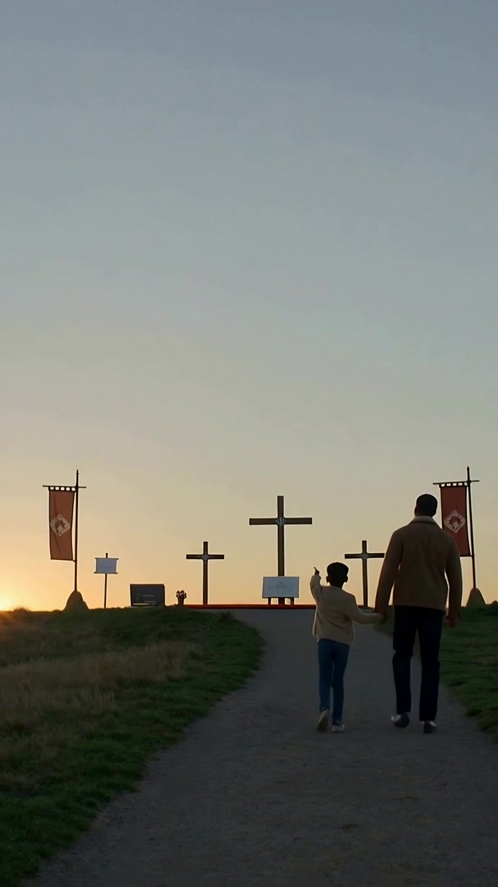 Video verticale: Alba che spinge un papà con cappotto e berretto ad accompagnare un bambino su per un sentiero verso le croci sulla collina per un memoriale.