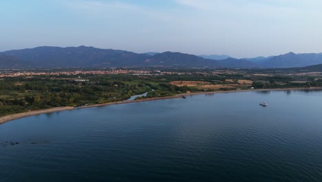 coastline of pula in sardinia at sunrise, yacht on calm sea, lush coast and distant mountains, landscape, italy. aerial backward, copy space
