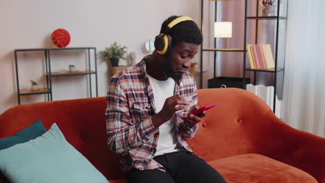 african american young man in headphones listening music dancing, singing in living room at home