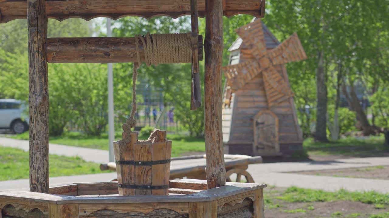 A wooden well with a bucket and a windmill in the background in the park.