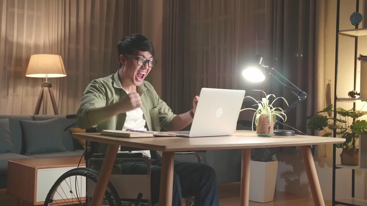 Asian Man Sitting In A Wheelchair While Working On Laptop Computer And Celebrating At Home, Night Time