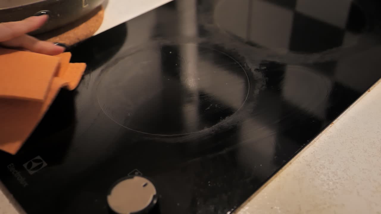 Woman cleaning electric stove with a spray cleaner and orange cloth, close-up.