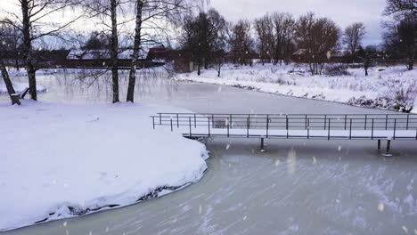 small city park with bridge over frozen lake water in winter season while snowing, aerial view