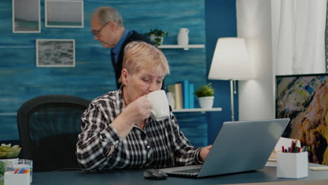 Older mature business woman using laptop sitting at workplace drinking coffee