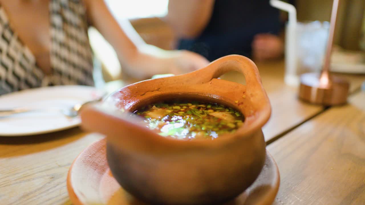 Hand lifts clay pot lid, revealing steaming herbal Thai soup in warmly lit Bangkok restaurant