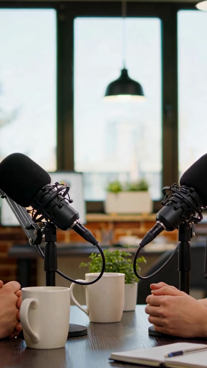 Video verticale: Registrazione di due persone che parlano e gesticolano a un tavolo vicino a una finestra, con microfoni per un podcast.
