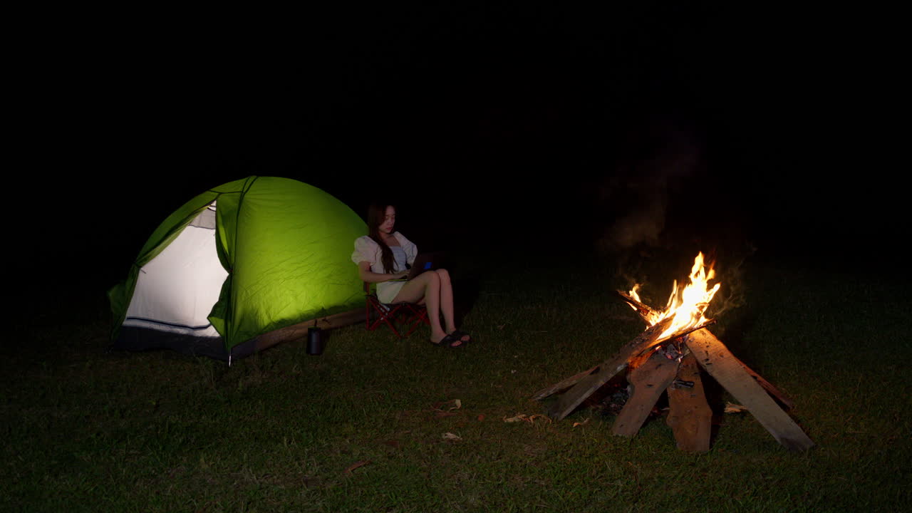 nómada digital trabajando remotamente concepto, joven asiática sentada con portátil fuera de su tienda delante de la hoguera por la noche