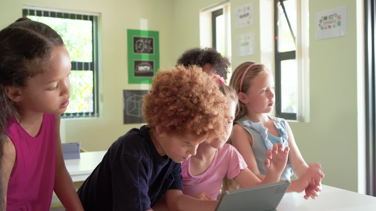 Diverse children clustering at classroom table while girl in pink tapping tablet to share screen