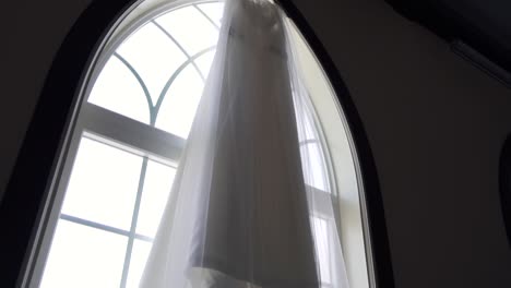 Upward-angled-push-in-shot-of-a-bride's-beautiful-wedding-dress-hanging-from-a-large-window-sill-waiting-to-be-worn-on-the-wedding-day