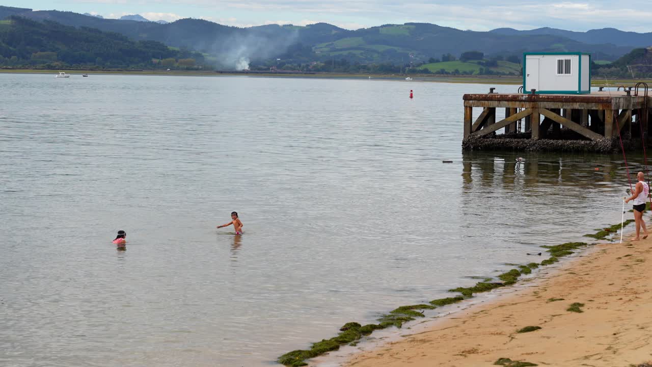 Kids swimming in the Cantabrian Sea as their father fishes from the beach in Santona, Cantabria