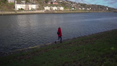 Slide-follow-motion-shot-of-a-girl-in-red-walking-on-a-river-bank-on-a-bright-and-sunny-day-in-nature-in-Ireland