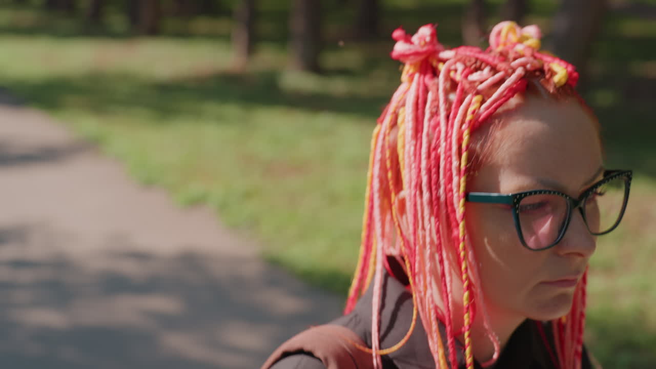 공원 주변을 즐기는 사려 깊은 여성, 화려한 땋은 머리를 한 여성이 빛나는 햇빛 아래 공원을 거닐고 있어요. 무지개색 땋은 머리를 한 여성이 화창한 공원을 생각에 잠겨 걸어가고 있어요.