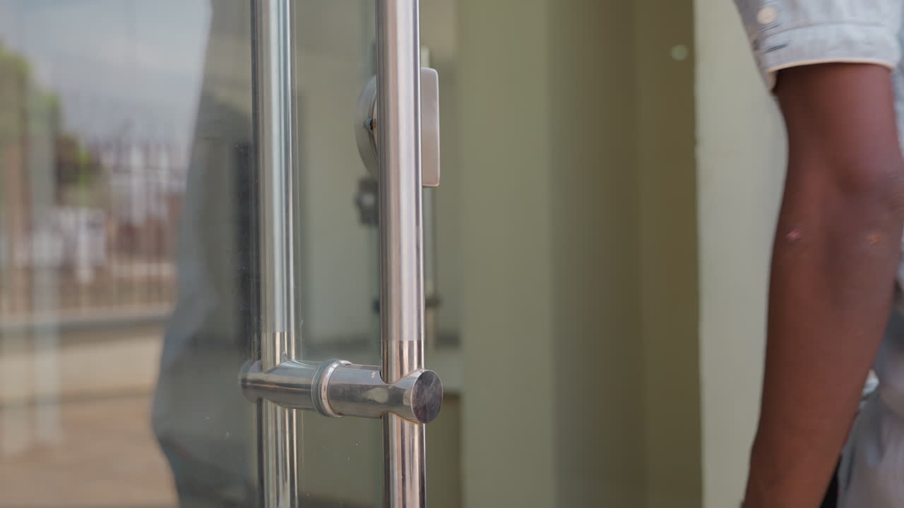 Tecnico nero che regola la cerniera della porta a vetri di una casa. Operaio di manutenzione che testa l'allineamento della maniglia e il chiavistello. Primo piano di raccordi metallici e movimento del braccio. Facciata residenziale e finestra riflettente.