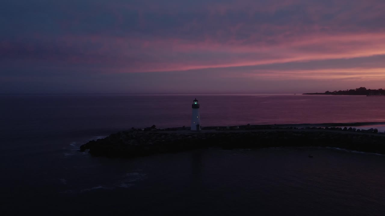 A drone shot circling the Walton Lighthouse, the small crafts harbor, and the beachfront in Santa Cruz California in front of a stunning sunset with waves crashing on the beach in the Pacific Ocean