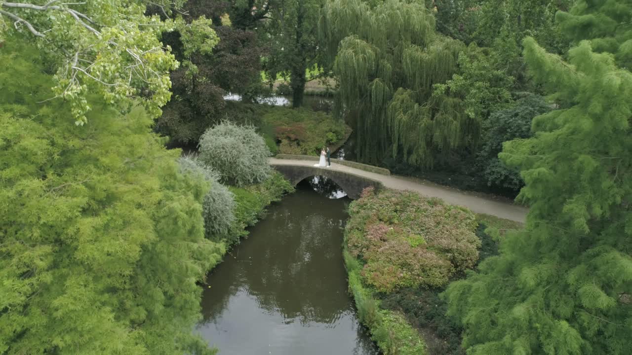 작은 다리 위에 서 있는 젊은 밀레니얼 웨딩 커플의 공중 돌리 아웃 슬로모션