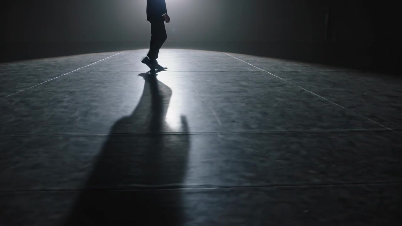 Entering round light pool, man in dark coat onstage crossing shiny stage floor, casting long shadow