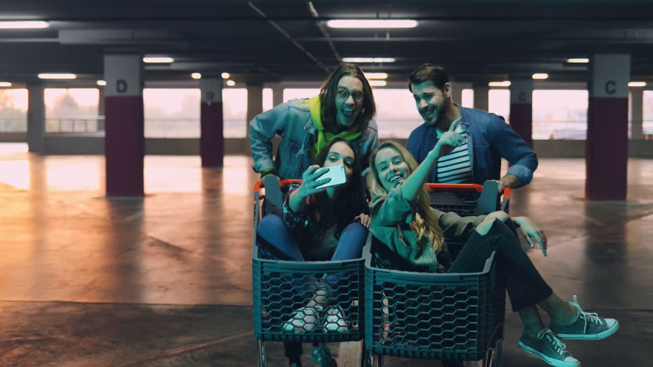 명랑한 젊은 친구들이 셀카를 찍거나 스마트폰으로 화상 통화를 하는 동안 소녀들은 트롤리에 앉아 있고 소년들은 그들을 짊어지고 있습니다