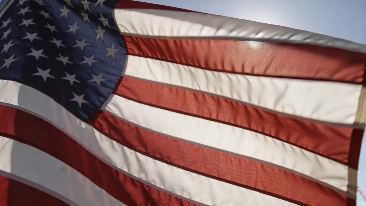 Grande bandiera americana che sventola, mostrando stelle e strisce sotto il sole splendente in un cielo blu, spinta dal vento.