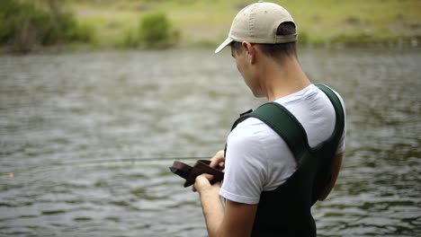 Toma-En-Cámara-Lenta-De-Un-Pescador-Masculino-Usando-Vadeadores-Preparando-Su-Cebo-Y-Mosca-Mientras-Pesca-Con-Mosca