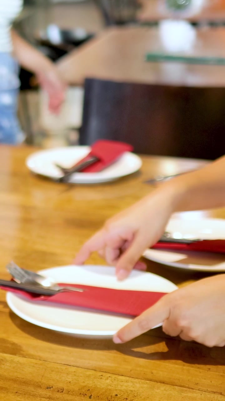 Hands arrange plates and utensils on a wooden table in a well-lit Bangkok restaurant