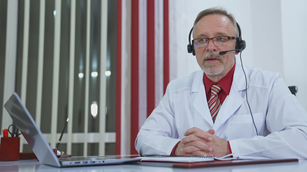 Doctor with headset talking on camera in medical office. Professional doctor in white medical coat sitting at desk and giving online consultation during quarantine.