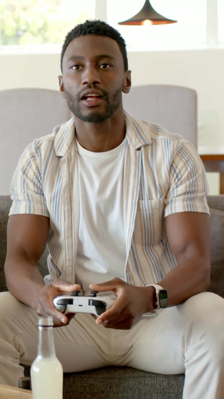 Vertical video: Putting bottle down, adult man grabbing controller and playing game in living room
