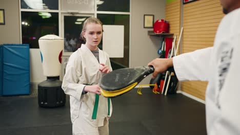 a young daughter woman wearing white robes does a technique taekwondo kung fu low kick into a padded cushion workout activity held by karate master inside a bright fitness center