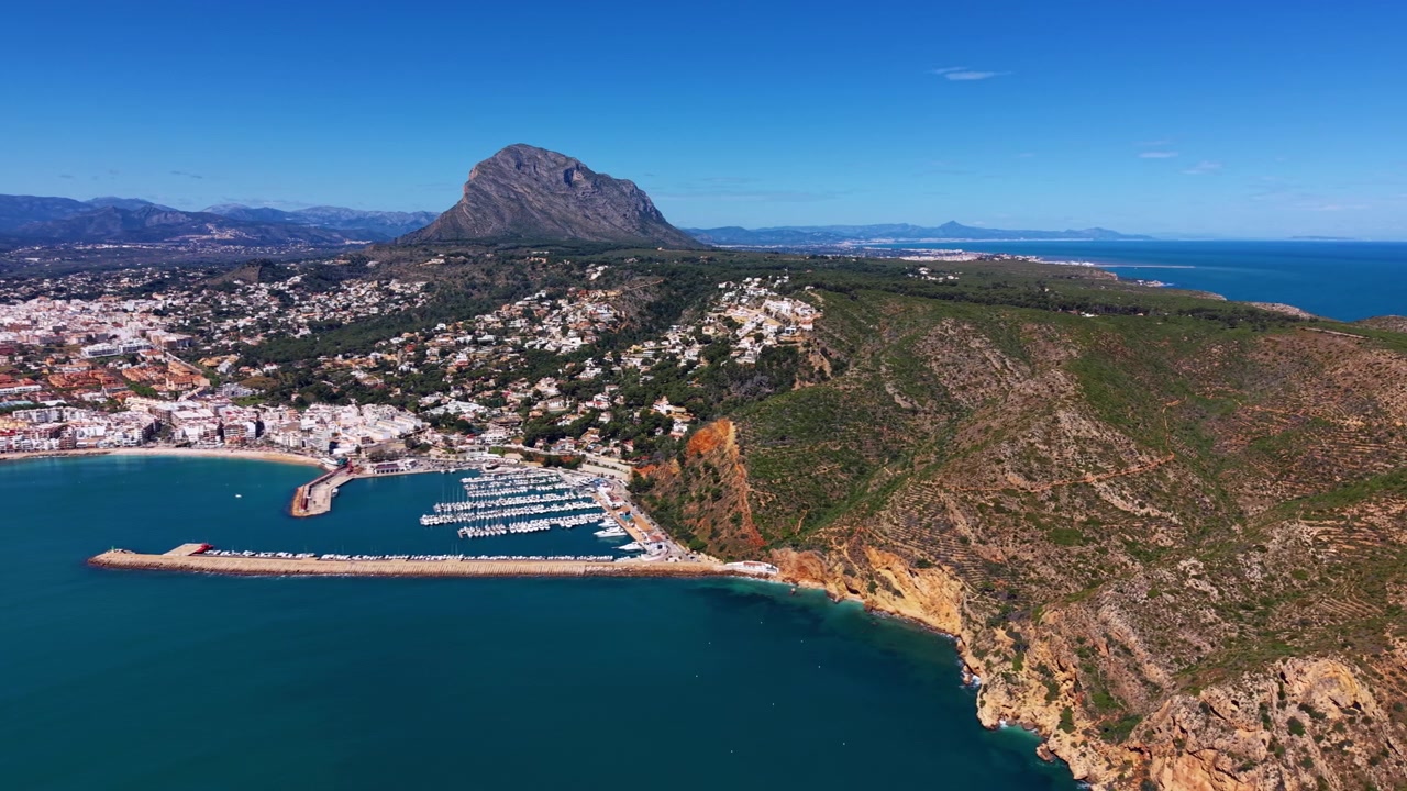 Città costiera di Javea sulla Costa Blanca, Spagna, con porto turistico, mare Mediterraneo turchese, scogliere rocciose, ville in collina e il monte Montgó sullo sfondo.