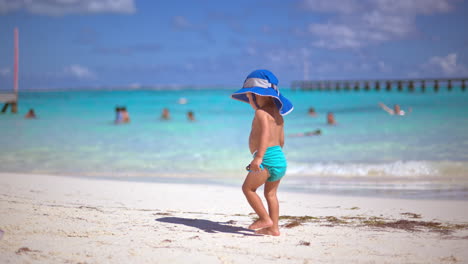 Niño-Pequeño-Con-Pañales-Turquesas-Y-Un-Sombrero-Azul-Parado-En-La-Playa-Mirando-El-Mar-Sosteniendo-Un-Auto-Azul-En-Cancún-México