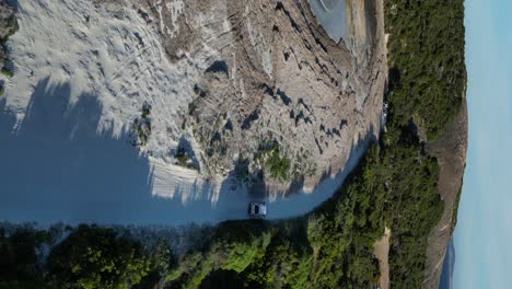 Vehicle-traveling-on-white-sand-across-trees-in-Wylie-Bay-Rock-Beach,-vertical-aerial