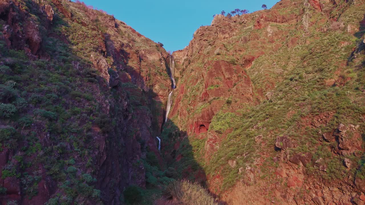 Veduta aerea della Cascata di Paul do Mar che scorre attraverso ripide scogliere vulcaniche rosse sull'isola di Madeira, circondata da terreno accidentato e luce solare calda che evidenzia il paesaggio drammatico