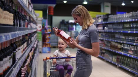 atractiva madre joven está eligiendo una botella de vino en el departamento de bebidas en el supermercado, mientras que su pequeño bebé es