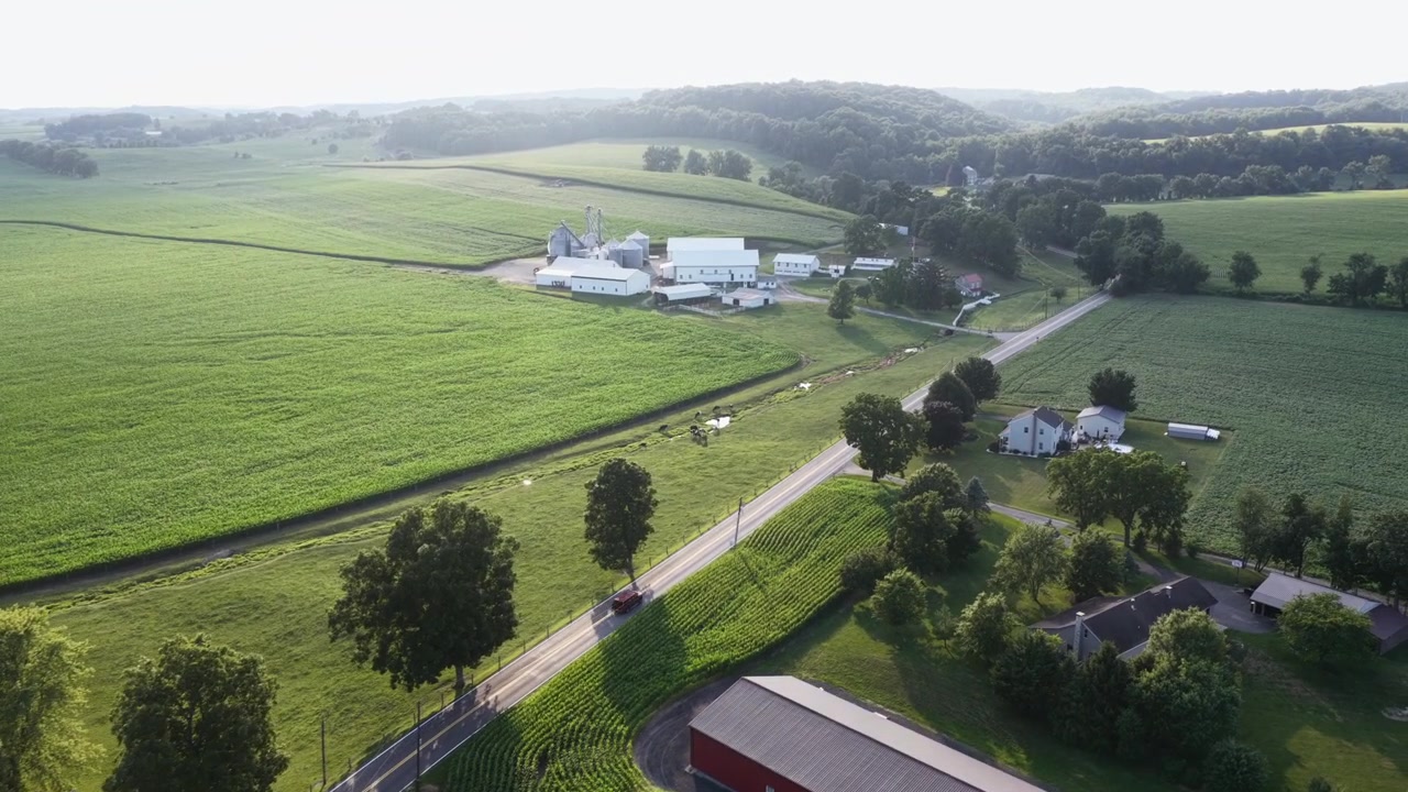 To-sporet vej bugter sig forbi grønne majsmarker og en rød metalstald, en bil kører ad landevejen mod fjerne hvide landbrugsbygninger og en bølgende skovklædt bakkeside i landlige USA. Luftfoto fra drone.