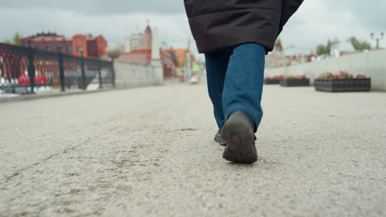 Nahaufnahme von Füßen, die auf einer Stadtpromenade entlanggehen, Low- Angle-Ansicht von Schuhen und Jeans, die strukturiertes Pflaster in der Nähe eines Brückengeländers und Flussuferarchitektur überqueren, gleichmäßiger Schritt, praktisches Schuhwerk, bedeckter Himmel